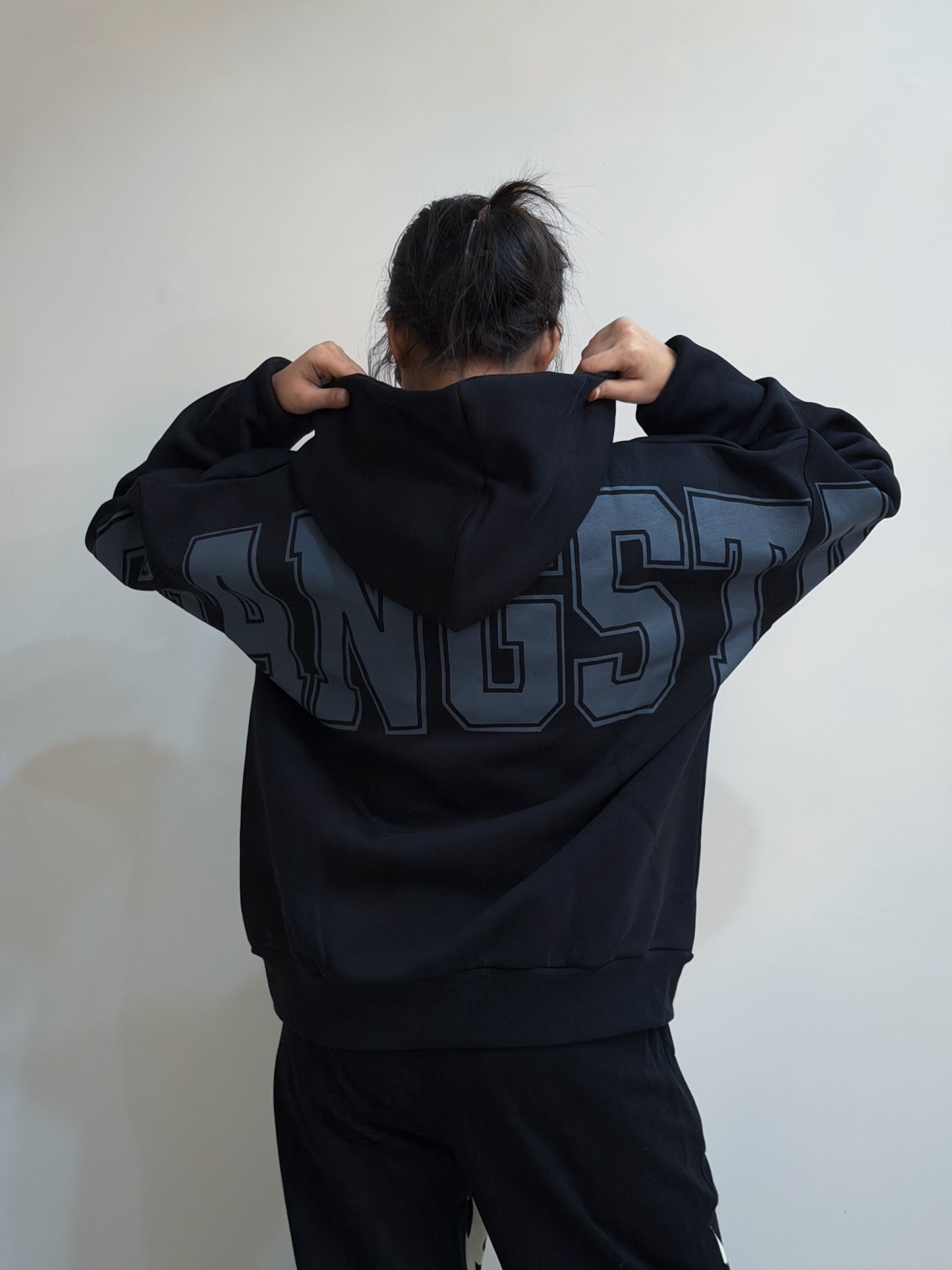 Person wearing a black hoodie with 'GANGSTA' printed on the back against a white wall