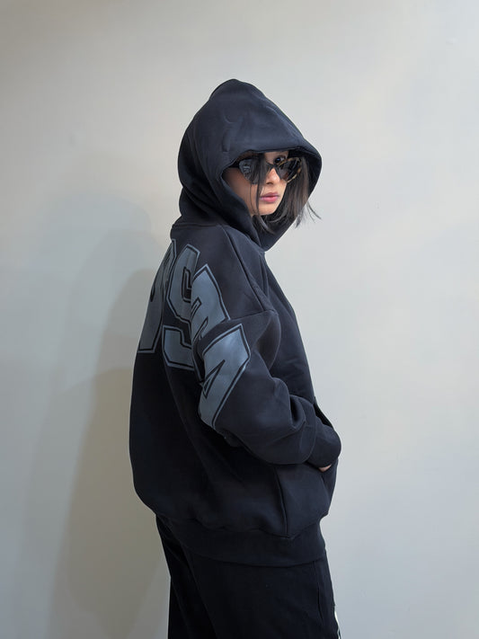 Person wearing a black hoodie with a logo on the sleeve against a plain background