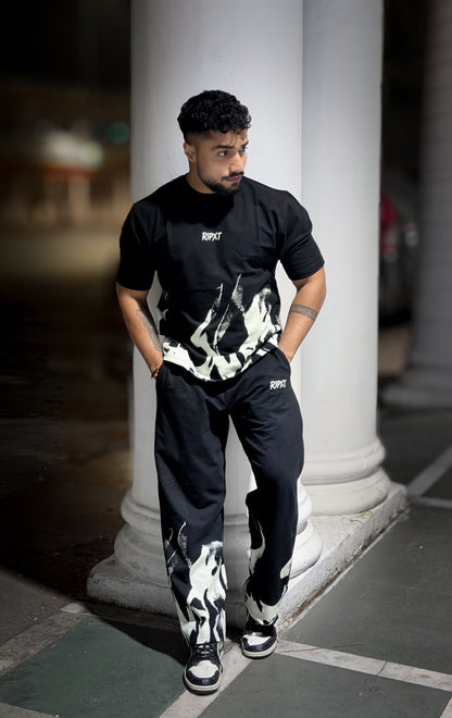 Man wearing a black t-shirt and pants with white patterns, standing against a blurred background.