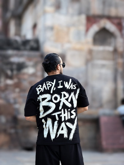 Person wearing a black t-shirt with white text "baby i was born this way" in an outdoor setting