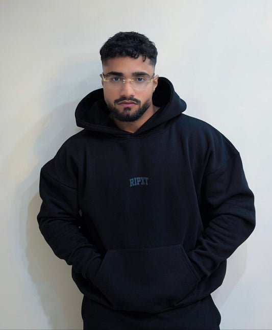 Man wearing a black hoodie with 'RIPXT' logo against a white wall