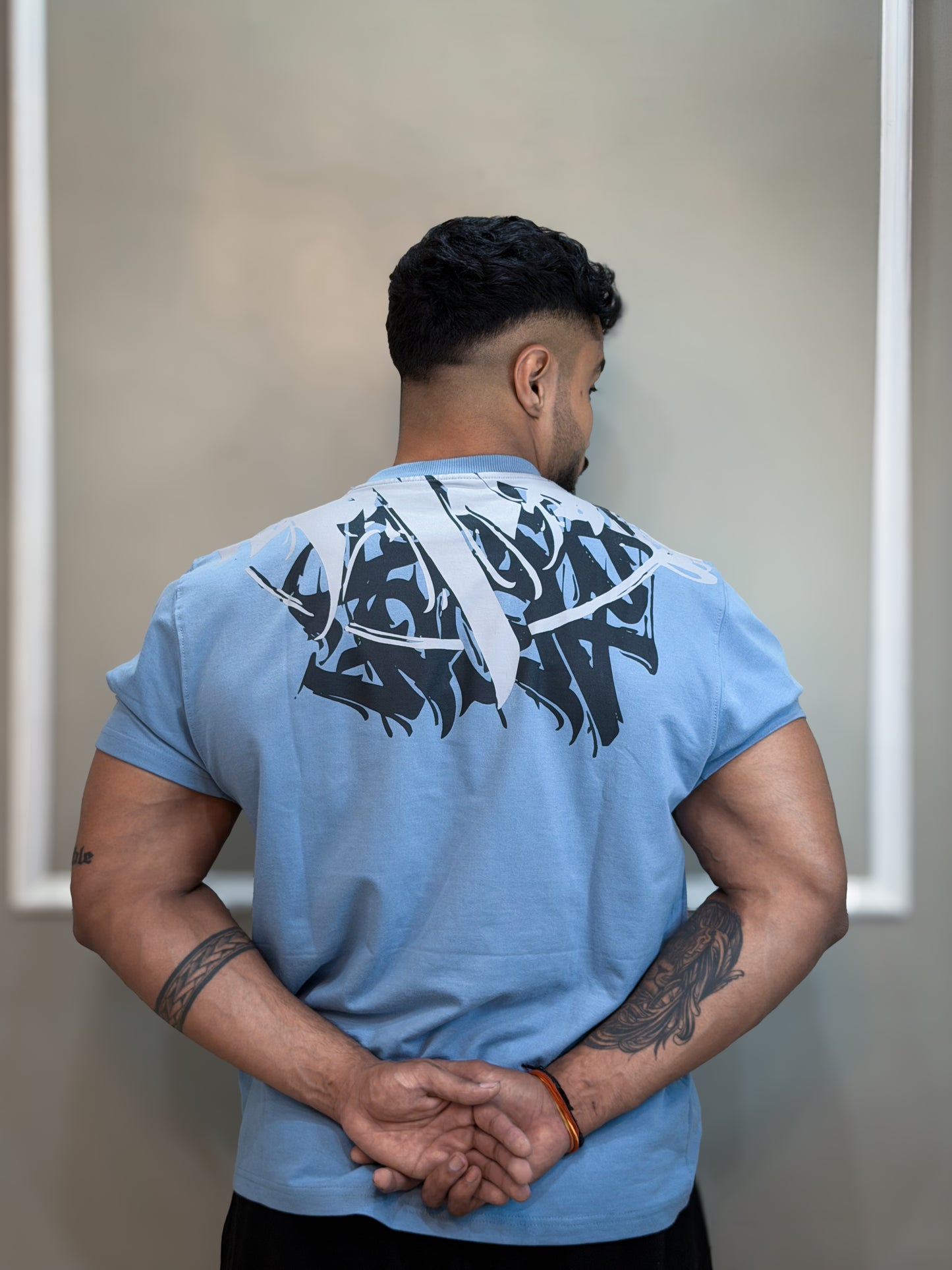 Man wearing a blue t-shirt with a black graphic design on the back, standing against a plain wall.